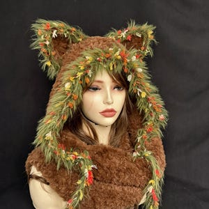 May include: A brown faux fur hood featuring two pointed ears and a decorative trim of green faux fur accented with small red, yellow, and white fabric flowers. The hood drapes around the neck and shoulders with long, dangling green fur tassels.