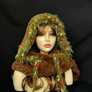 May include: A brown faux fur hooded cowl featuring a decorative trim of green faux grass fibres interspersed with small yellow, orange, and white floral accents. The hood frames the face, with two long, thin green fibre strands hanging down.