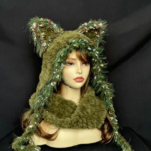 May include: An olive green faux fur hood featuring two large pointed ears. The hood is trimmed with long, textured green and white fringe along the edges and includes two long, dangling side panels that drape down from the main hood structure.