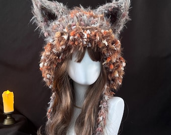 Puede incluir: Un gorro texturizado y peludo con orejas de animal, hecho de hilo jaspeado en tonos marrón, gris, blanco y naranja. El gorro presenta orejas puntiagudas y prominentes en la parte superior y paneles laterales largos que cuelgan y enmarcan el rostro.
