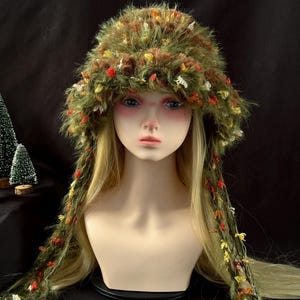May include: A fuzzy, olive green bucket hat with long, dangling side panels. The hat is textured with integrated, multi-colored yarn accents in shades of red, orange, and yellow, creating a dense, shaggy appearance that covers the head and shoulders.