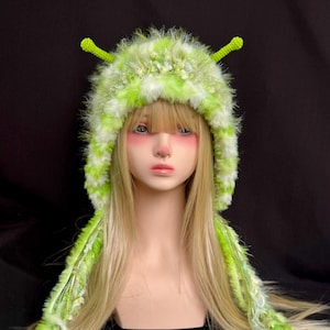 May include: A fuzzy, lime green and white variegated yarn hat featuring two upright, textured green antennae on top. The hat has long, dangling side panels made of the same shaggy, multi-textured yarn, creating a soft, layered appearance.