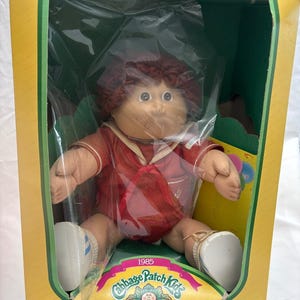 Könnte beinhalten: Eine Cabbage Patch Kids Puppe aus dem Jahr 1985 mit rotbraunem Wollhaar, bekleidet mit einem roten Matrosenoutfit mit weißem Kragen. Die Puppe befindet sich in einem gelb-grünen Karton mit dem Text Cabbage Patch Kids und I'm unique. I'm adoptable.
