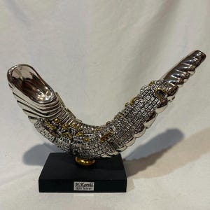 May include: A silver-colored shofar sculpture featuring intricate city wall detailing with gold-toned accents, mounted on a rectangular black base. A small white label on the base reads H.Karshi 925 silver. The object has a curved, horn-like shape.