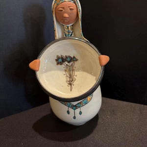 May include: A ceramic figurine of a person wearing a head covering, holding a round bowl with a dark rim. The bowl features a painted floral design in brown and blue. The figurine has a cream-colored base with blue and gold geometric accents.