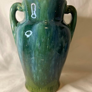 Può includere: Un vaso in ceramica caratterizzato da una smaltatura lucida e screziata nei toni del verde bosco profondo e del verde acqua. Il vaso presenta un'apertura ampia, un corpo affusolato e due manici simmetrici e curvi sui lati superiori.