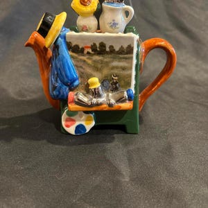 Puede incluir: Una tetera de cerámica con forma de caballete de artista, que presenta un paisaje pintado, un abrigo azul con un sombrero amarillo, una paleta de pintura y jarrones en miniatura con flores y pinceles en la parte superior. El asa y el pico son de color marrón anaranjado brillante.