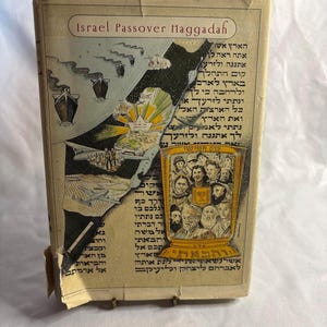 以下が含まれることがあります： ベージュ色の表紙の「Israel Passover Haggadah」というタイトルの古い本。表紙にはイスラエルの地図、船や飛行機のイラスト、ヘブライ語のテキスト、そして人々のグループとメノーラーが描かれた金色のグラフィックが配置されています。