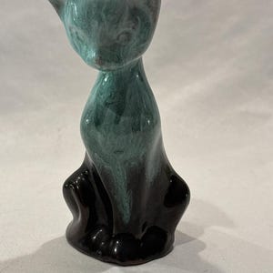 May include: A ceramic cat figurine featuring a glossy finish with a color gradient that transitions from a deep, solid black at the base to a mottled, swirling teal and dark green pattern across the upper body, head, and pointed ears.
