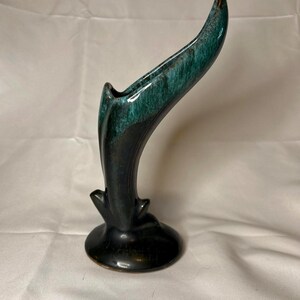 May include: A ceramic vase with a flared, asymmetrical opening and a curved, elongated silhouette. The piece features a deep black base that transitions into a mottled, vibrant teal glaze at the top, with a smooth, glossy finish throughout.
