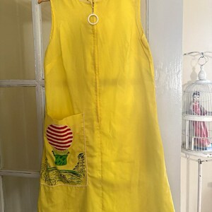 May include: A sleeveless yellow dress featuring a front zipper closure with a white circular pull tab. A patch pocket on the lower front displays a graphic illustration of a red and white striped hot air balloon above a landscape scene.