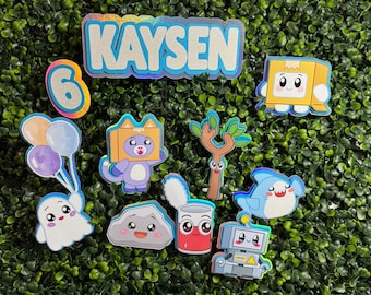 May include: A set of cake toppers featuring the name KAYSEN in silver glitter with blue borders, the number 6, and various cartoon characters including a ghost with balloons, a cat in a box, a tree, a rock, a can, a shark, and a robot.