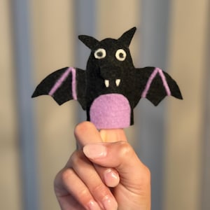 Può includere: Un burattino da dito in feltro nero a forma di pipistrello con pancia viola, occhi tondi bianchi con pupille nere e due piccole zanne bianche. Le ali presentano dettagli cuciti in viola e il burattino è realizzato in feltro stratificato.