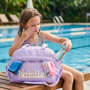 Peut inclure: Un sac de sport violet clair avec le prénom Natalie brodé en écriture blanche sur la poche avant. Le sac contient un tissu bleu plié, une paire de lunettes de natation grises et une bouteille d'eau transparente dans le compartiment latéral.