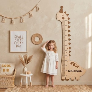 May include: A wooden wall-mounted height chart shaped like a long-necked dinosaur with a party hat. The chart features black measurement markings from 70 to 180 centimeters and the name MADISON printed in black on the dinosaur's body.