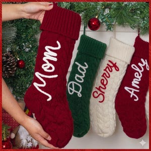 May include: Four cable-knit Christmas stockings with folded cuffs. The first is deep red with white script reading Mom, the second is dark green with white script reading Dad, the third is cream with red script reading Sherry, and the fourth is red with white script reading Amely.