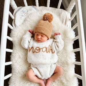 May include: A baby wearing a tan ribbed knit beanie with a pom-pom and a cream-coloured speckled knit sweater and nappy cover set. The sweater features the name Noah embroidered in tan yarn. The baby is lying in a white wooden crib with a nameplate.