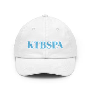 Op de afbeelding: Witte baseballpet met een gebogen klep. De pet heeft de lichtblauwe tekst "KTBSPA" op de voorkant. De pet heeft een knoop bovenop en een verstelbare band aan de achterkant.