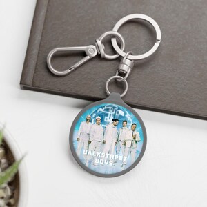 Pode incluir: Um porta-chaves dos Backstreet Boys com uma foto de grupo em roupas brancas sobre um fundo azul. O porta-chaves tem uma borda cinza e ferragens prateadas, incluindo um mosquetão e um porta-chaves. O texto "BACKSTREET BOYS" é visível.