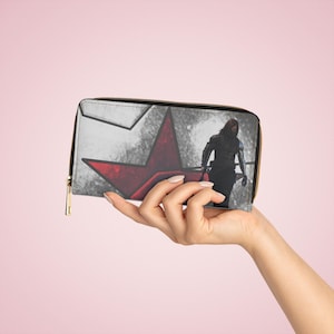 May include: A black and white wallet with a red star design and a character illustration. The wallet has a gold zipper and is held by a hand against a pink background. The wallet is approximately 8 inches long.