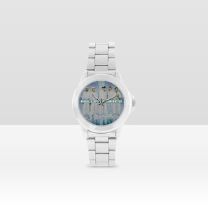 May include: A silver-toned wristwatch with a band and face featuring a group of people in white outfits. The watch face has the text "MILLA JUM 20" in a light blue color.