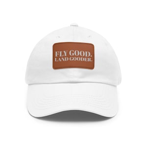 Puede incluir: Gorra de béisbol blanca con un parche rectangular marrón. El parche presenta el texto "FLY GOOD. LAND GOODER." en color marrón claro. La gorra tiene una visera curva y un diseño clásico e informal.
