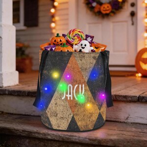 Può includere: Un secchiello per dolci di Halloween cilindrico con un motivo a rombi nero e marrone e il nome JACK stampato in bianco. Il secchiello è illuminato da piccole luci LED colorate ed è riempito con vari dolci, un lecca-lecca e zucche.