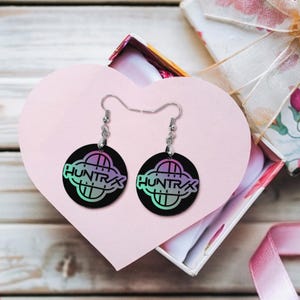May include: A pair of black round earrings with the word "HUNTRX" in a colorful gradient design. The earrings are displayed in a heart-shaped box, suggesting a gift.