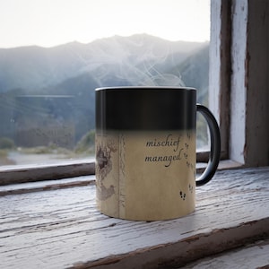 Può includere: Una tazza nera termosensibile con le parole "mischief managed" in un carattere script. La tazza presenta un disegno a mappa e impronte di zampe. La tazza è appoggiata su un davanzale di legno bianco e usurato con vista sulle montagne sullo sfondo.