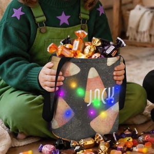 Può includere: Una borsa a secchiello in tessuto nero con un motivo a caramelle di mais e il nome Jack stampato in bianco. La borsa è illuminata da piccole luci LED colorate ed è riempita con vari dolci incartati, inclusi involucri in lamina arancione e oro.