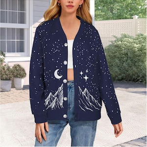 May include: Navy blue cardigan with a celestial design. The jacket features a white mountain range, crescent moon, stars, and button closure. The long-sleeved cardigan is a comfortable outerwear option.