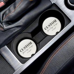 May include: Two circular white car cup holder coasters featuring the black printed text CUSTOM Your logo, image, or text! The coasters are positioned inside the black plastic cup holders of a vehicle center console with leather trim.