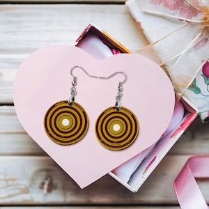 May include: A pair of round earrings with a target design in shades of gold, maroon, and white. The earrings are suspended from silver-colored hooks. The earrings are presented on a pink heart-shaped card inside a gift box.