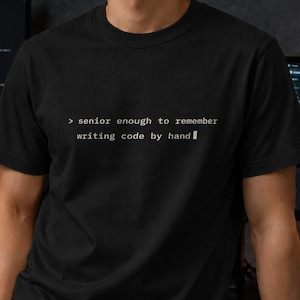 May include: A black short-sleeved t-shirt featuring white text on the chest that reads: > senior enough to remember writing code by hand. The shirt has a crew neckline and a standard fit, constructed from a solid black fabric with white lettering.