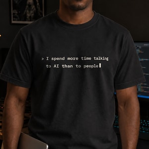 Puede incluir: Una camiseta negra de manga corta y cuello redondo con texto blanco impreso en el pecho que dice: > I spend more time talking to AI than to people. La prenda tiene un corte estándar con cuello acanalado y está hecha de tela oscura.