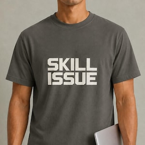 May include: A charcoal grey short-sleeved t-shirt featuring the text SKILL ISSUE printed in a bold, white, block-style font across the chest. The garment has a crew neckline and a standard fit, providing a clean and straightforward appearance.