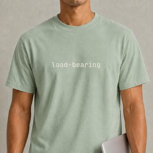 May include: A sage green short-sleeved t-shirt featuring the text "load-bearing" printed in white across the chest. The shirt has a crew neckline and a standard fit, paired with blue denim jeans while holding a silver laptop computer.