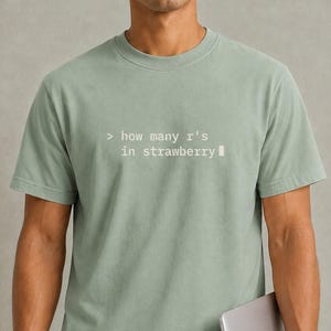 May include: A sage green short-sleeved t-shirt featuring white monospaced text on the chest that reads > how many r's in strawberry with a blinking cursor block at the end of the line. The shirt has a crew neckline and a standard fit.