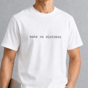 May include: A white short-sleeved t-shirt featuring the text "make no mistakes" printed in a simple black sans-serif font across the chest. The garment has a classic crew neckline and a straight hemline, providing a clean and basic appearance.