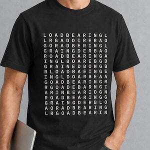 Puede incluir: Una camiseta negra de manga corta con un gran gráfico cuadrado blanco en el pecho. El gráfico consiste en una densa cuadrícula de letras que deletrean la frase LOAD BEARING repetidamente en varias configuraciones a lo largo de quince filas.