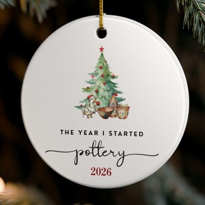 May include: A round white ceramic ornament hanging by a gold string. The centre features a watercolour illustration of a Christmas tree, a goose, and pottery items. Below the image, the text reads THE YEAR I STARTED pottery 2026.