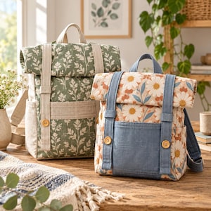 May include: Two roll-top backpacks made of fabric. The left backpack features a green background with a white fern leaf pattern and beige straps. The right backpack features a white background with orange and white daisy flowers and blue straps.