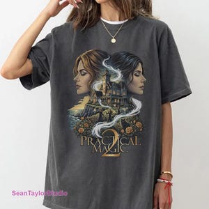 May include: A charcoal gray short-sleeve t-shirt featuring a graphic print of two women in profile flanking a large house on a cliffside. Below the house are figures, roses, and a black cat. The text Practical Magic 2 is printed in gold lettering.