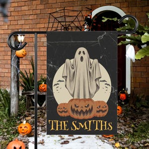 Könnte beinhalten: Eine rechteckige Gartenflagge mit schwarzem Hintergrund, einem zentralen beigen Mond, einem weißen Geist und drei orangefarbenen Kürbissen. Der Text THE SMITHS ist unten in Gelb aufgedruckt. Die Flagge hängt an einem schwarzen Metallständer.