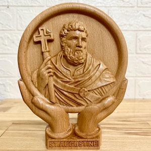 Op de afbeelding: Een lichtbruin houten snijwerk van de heilige Augustinus in een cirkelvormig frame dat door twee handen wordt vastgehouden. De figuur heeft een baard, draagt gewaden en houdt een kruis vast. De voet bevat de tekst ST. AUGUSTINE.