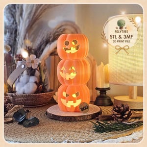 May include: A stack of three orange 3D printed pumpkins with carved jack-o'-lantern faces. The pumpkins are illuminated from within, casting a warm glow. A small circular tag with the word BOO is attached to the bottom pumpkin.