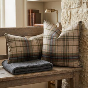 May include: Two square throw pillows featuring a beige, olive green, and blue plaid pattern. The pillows are arranged on a wooden bench alongside a folded dark gray knit throw blanket, creating a coordinated set of home textile accessories.