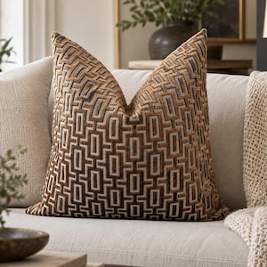 May include: A square throw pillow featuring a raised, textured geometric pattern of interlocking rectangles in shades of metallic bronze, copper, and light gray. The fabric has a velvet-like appearance with a distinct, repeating grid design.