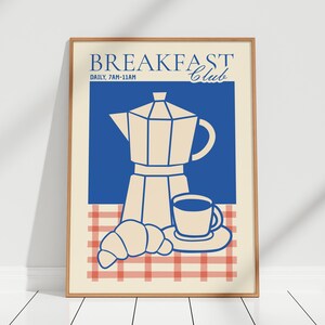 May include: A framed wall art print featuring the text BREAKFAST Club and DAILY, 7AM-11AM. The illustration shows a beige moka pot, a coffee cup on a saucer, and a croissant set against a blue background and a red and white checkered tablecloth.