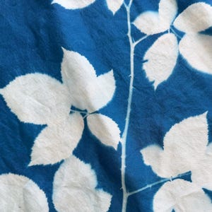 May include: A piece of fabric featuring a cyanotype print with a deep blue background and white silhouettes of leaves and stems. The pattern displays a botanical arrangement of foliage with jagged edges, creating a high-contrast visual effect.
